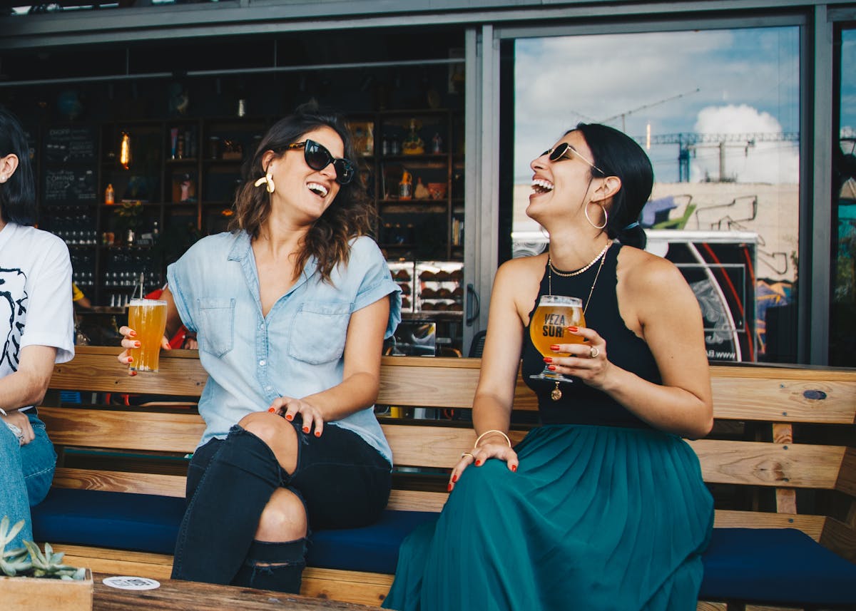 Two women laughing with beers on a bench